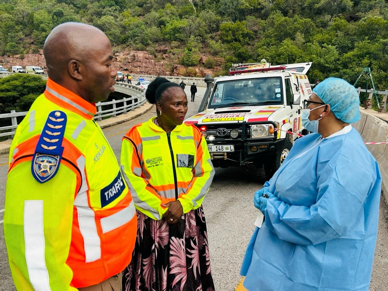 Emergency teams on the bridge where the bus crashed. Two are wearing high-viz uniforms. One is wearing pale blue medical overalls, a hat and gloves. There is a truck behind them.