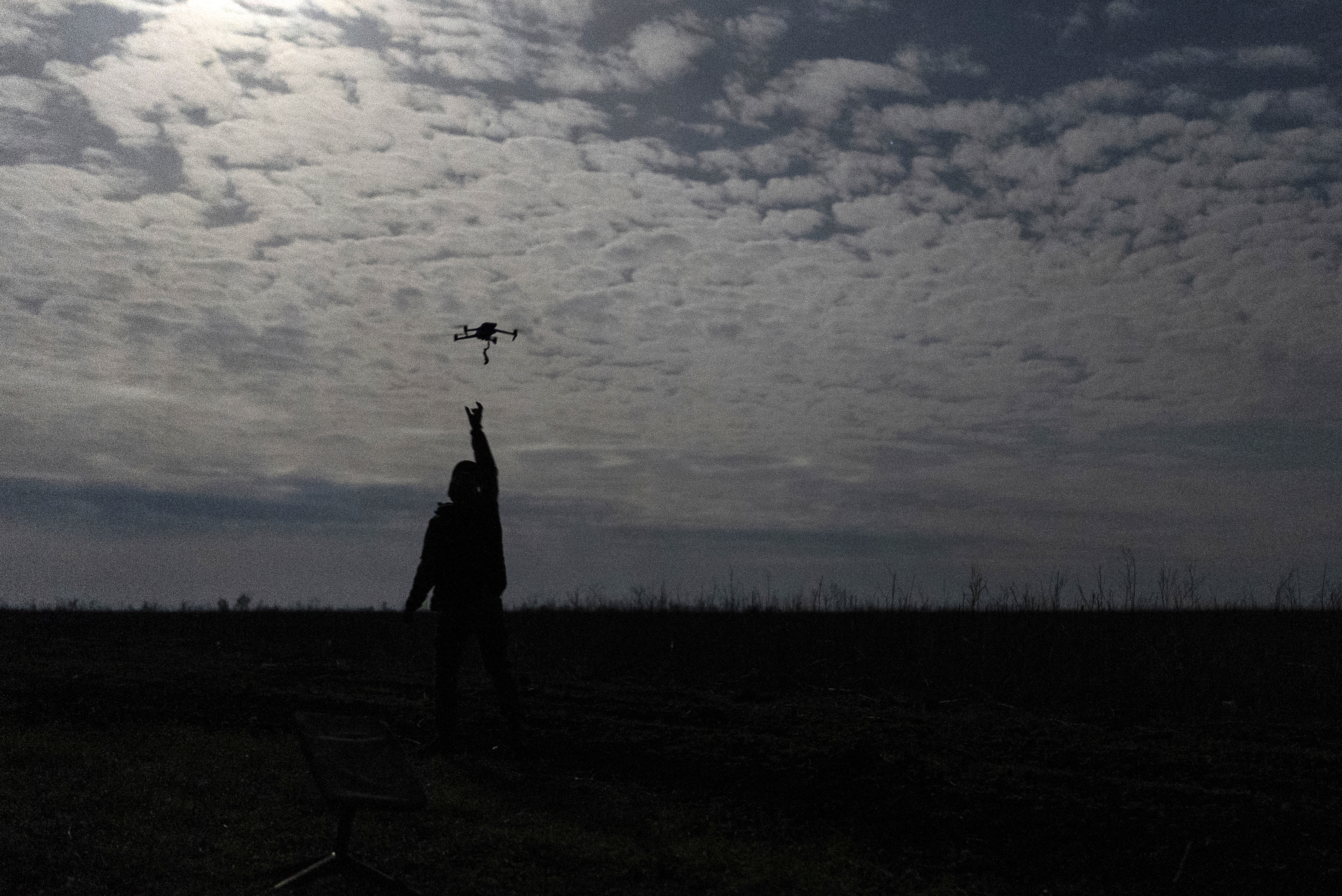 A Ukrainian soldier catches a drone on the eastern frontline. he is silhouetted against the sky and has his arm outstretched. The terrain in flat.